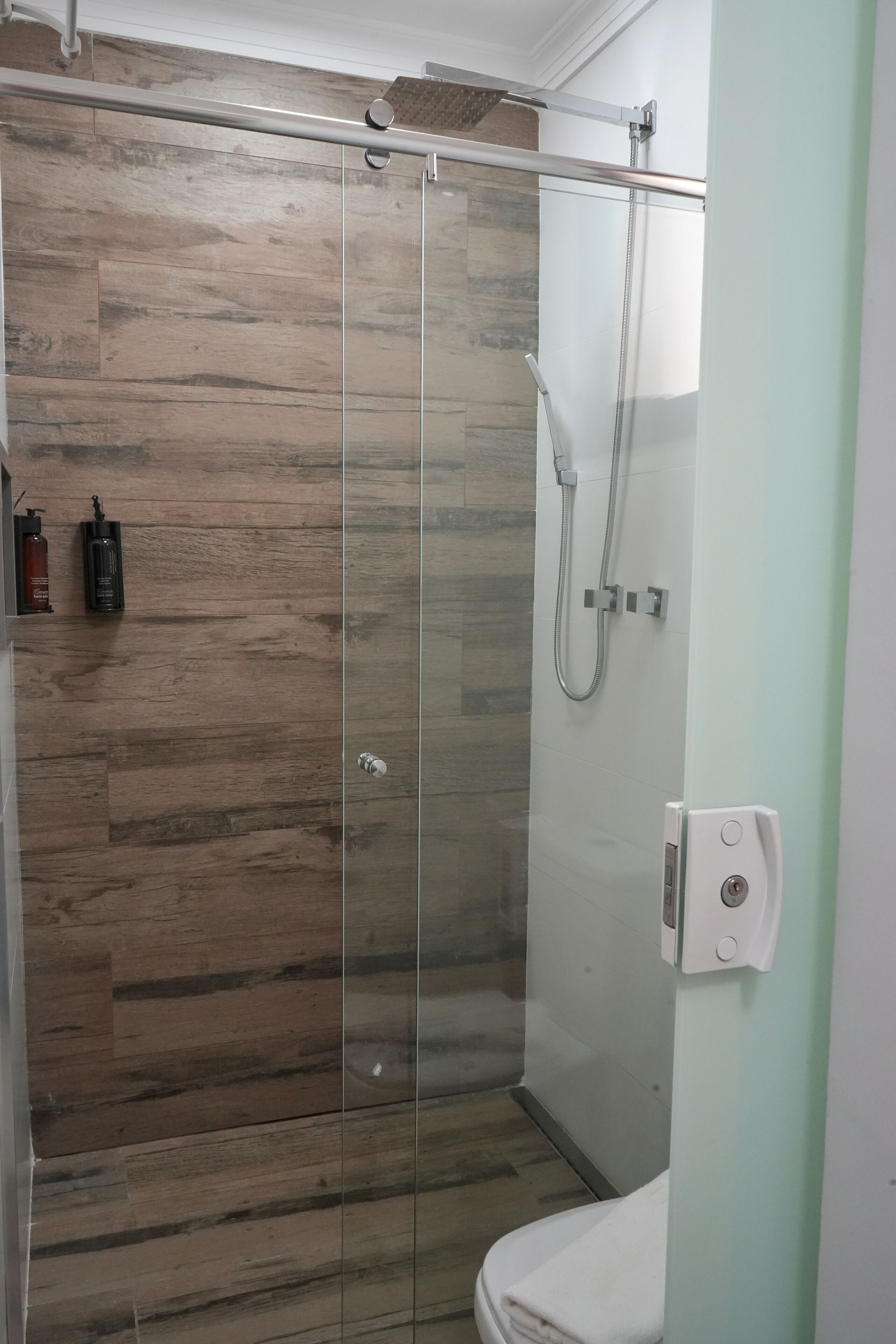 Una cabina doccia in vetro, pavimento in legno, un water e un dispenser di sapone a parete.