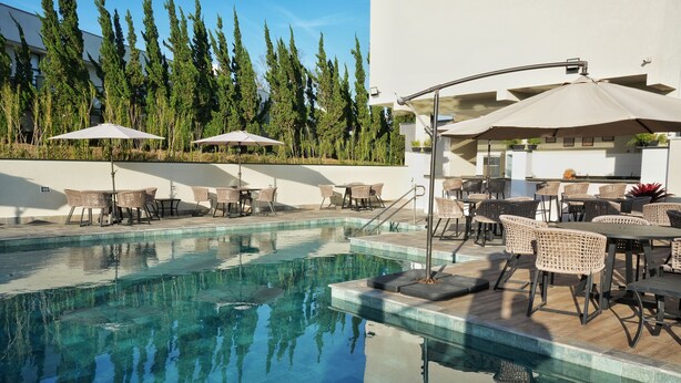 A pool area with lounge chairs and umbrellas, surrounded by tall trees and a building with a bar. - Bristol Villaggio Rosa Arujá (Arujá)