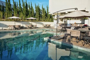 Uma área de piscina com espreguiçadeiras e guarda-sóis, cercada por árvores altas e um edifício com bar.