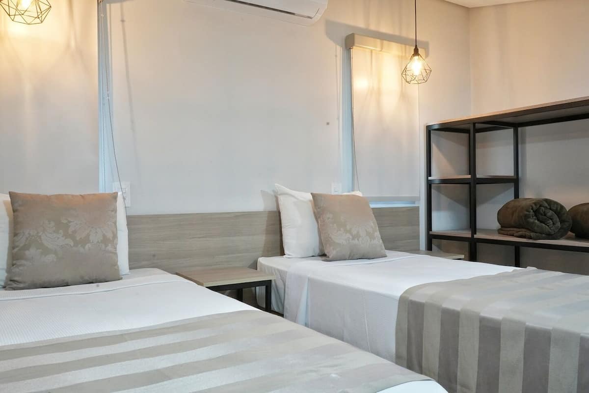 Habitación de hotel con dos camas, cabecera de madera, estante con mantas e iluminación colgante.