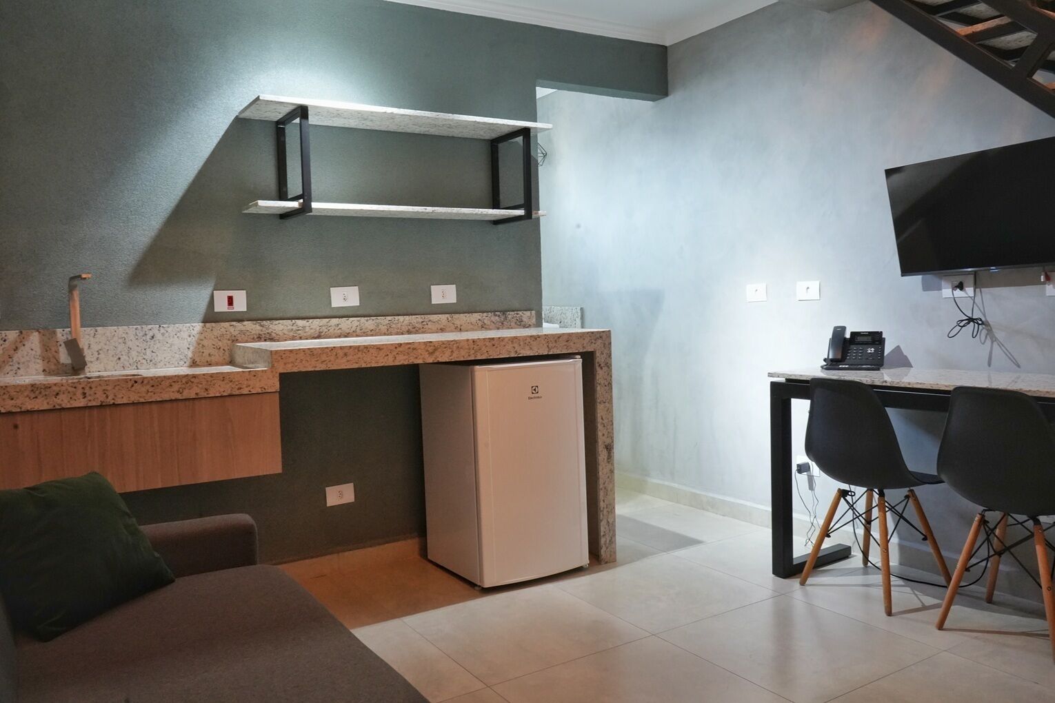 Una kitchenette compacta con refrigerador integrado, un lavamanos y una encimera. Hay una pequeña zona de estar con un sofá y un televisor montado en la pared.