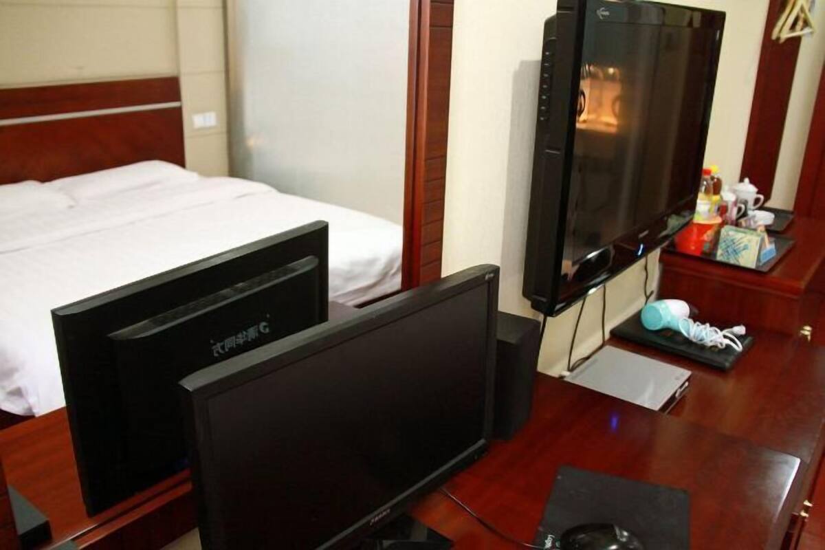 A hotel room with a bed, a television, a desk with a computer, and a small table with a cup and a box.