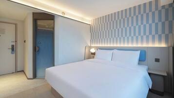 A hotel room with a large bed, a nightstand, a wall-mounted lamp, and a door leading to another room.