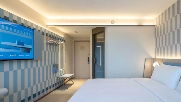 A modern hotel room with a bed, a TV mounted on the wall, a door, and a small table.
