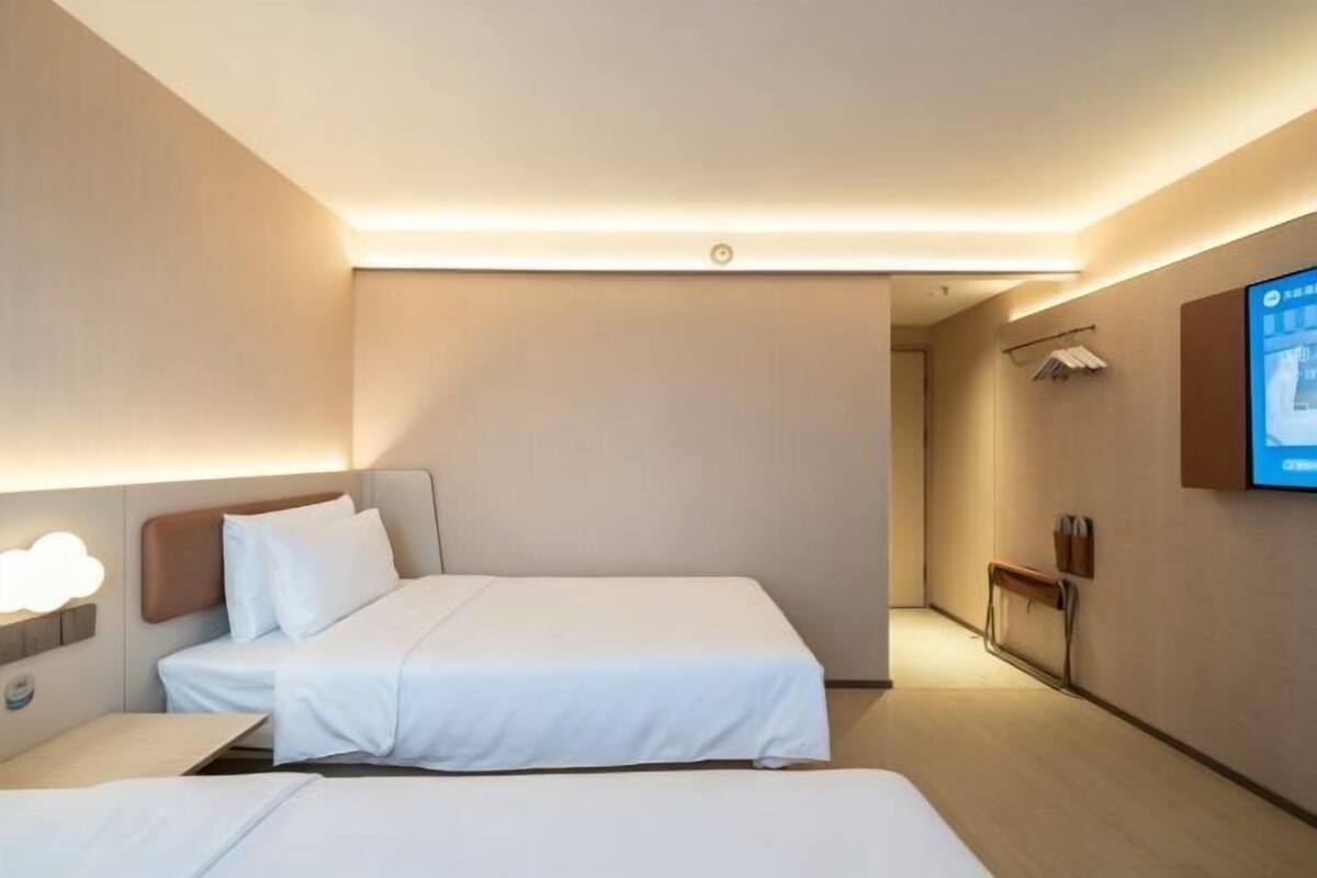 A hotel room with two beds, a TV mounted on the wall, and a bedside table with a lamp.
