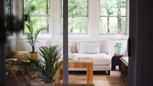 Une pièce avec un canapé blanc, une table basse en osier et des plantes en pot. De grandes fenêtres laissent entrer la lumière naturelle.
