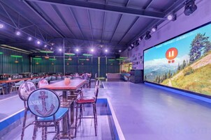 Un salón de conferencias con una pantalla grande mostrando un paisaje pintoresco, varias mesas con sillas y una zona de escenario.