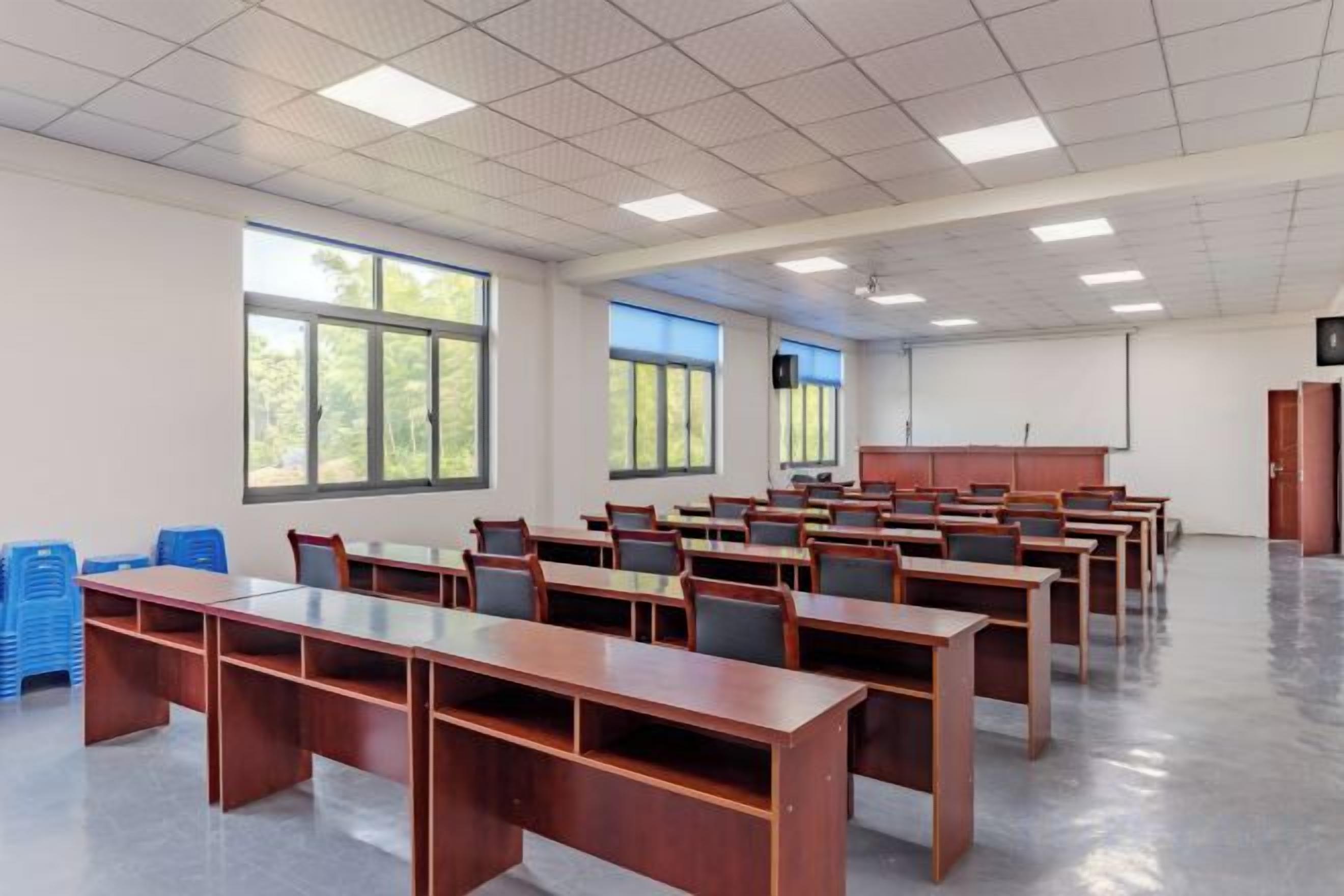 Une salle de classe avec des rangées de bureaux, de grandes fenêtres et un écran de projection.