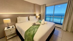 Hotelkamer met een groot bed, twee bedlampen, een nachtkastje met een afstandsbediening, een dienblad met een beker en een vaas, en uitzicht op de oceaan door een groot raam.