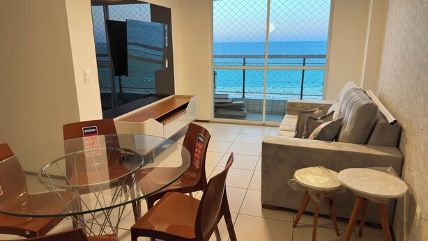 Een moderne woonkamer met een glazen eettafel, bruine stoelen, een bank en een televisie. Door grote ramen is een balkon met uitzicht op zee zichtbaar.
