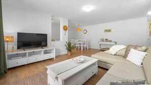 A living room with a TV, white furniture, a dining table, and a sofa. - Shelly Cove Apartment 12 - aircon, pool, beach across road (Port Macquarie)