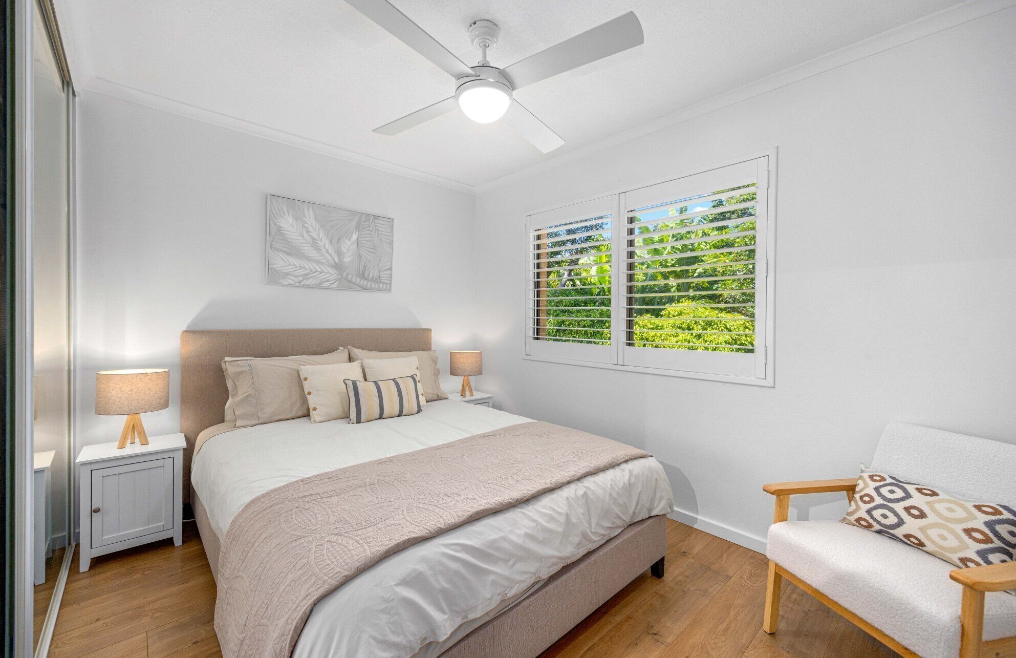 Une chambre à coucher comprenant un lit, des tables de chevet, une chaise, un ventilateur de plafond et une fenêtre donnant sur un paysage verdoyant.