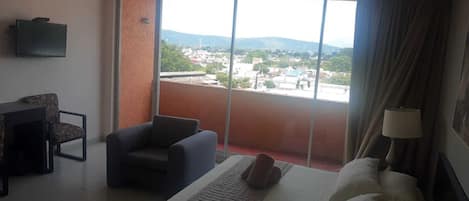 Habitación de hotel con cama, una silla, escritorio, televisor y ventana grande con vista a la ciudad.