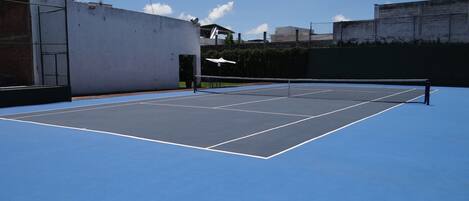 Una cancha de tenis exterior con superficie azul, rodeada de muros y una valla.
