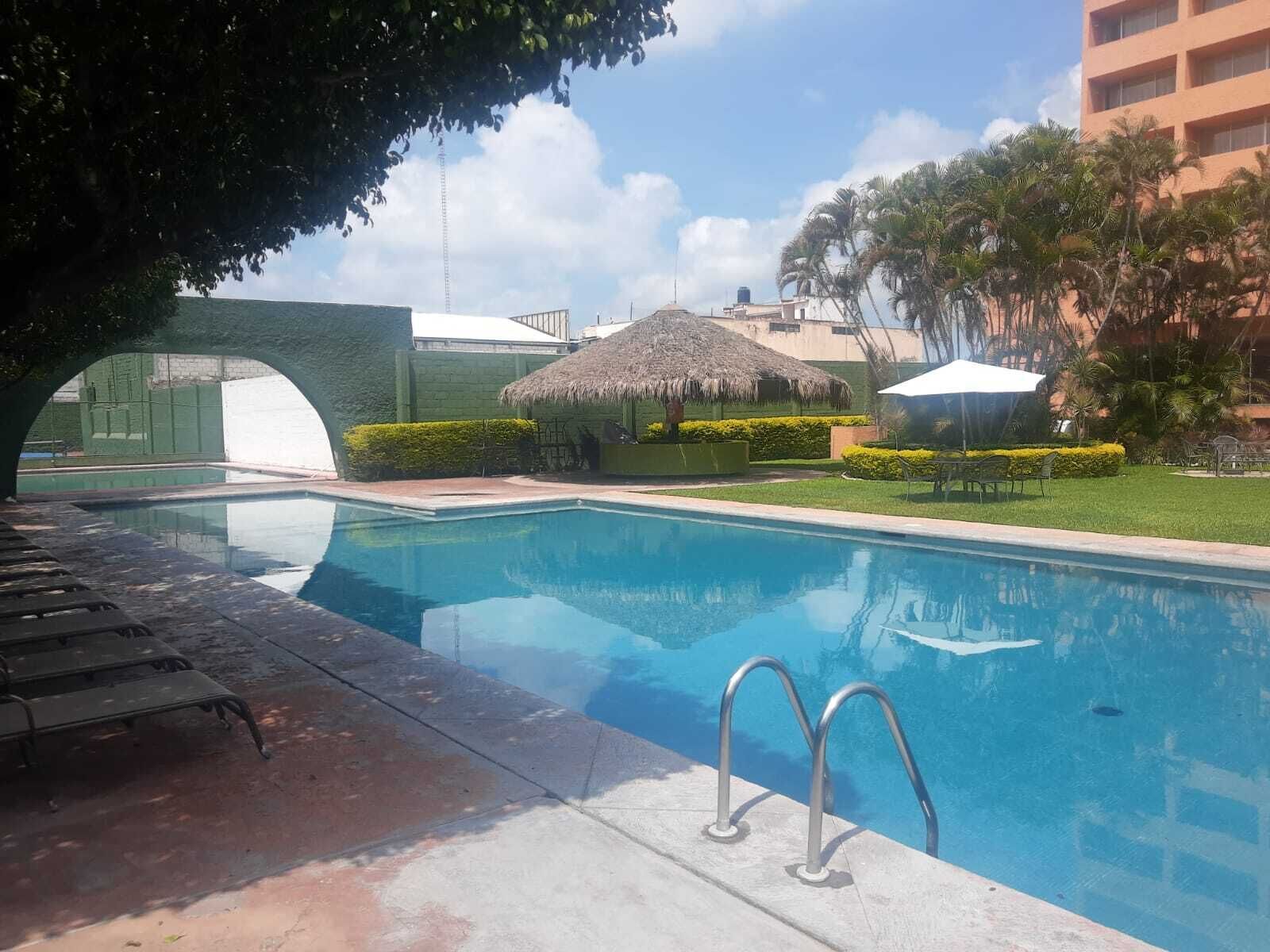 En swimmingpool med en stråtækt bygning, et overdækket siddeområde og udsigt til en bygning i baggrunden.