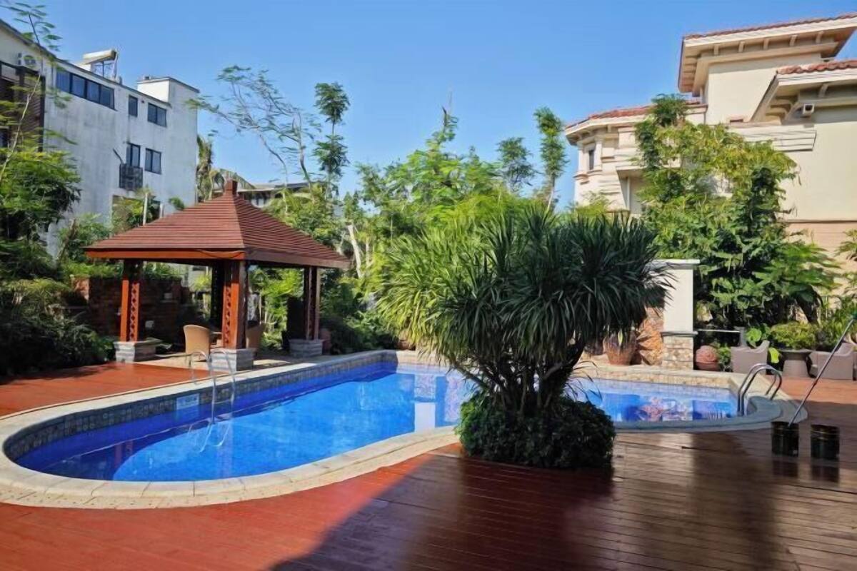 A swimming pool with a gazebo, surrounded by greenery and modern buildings.