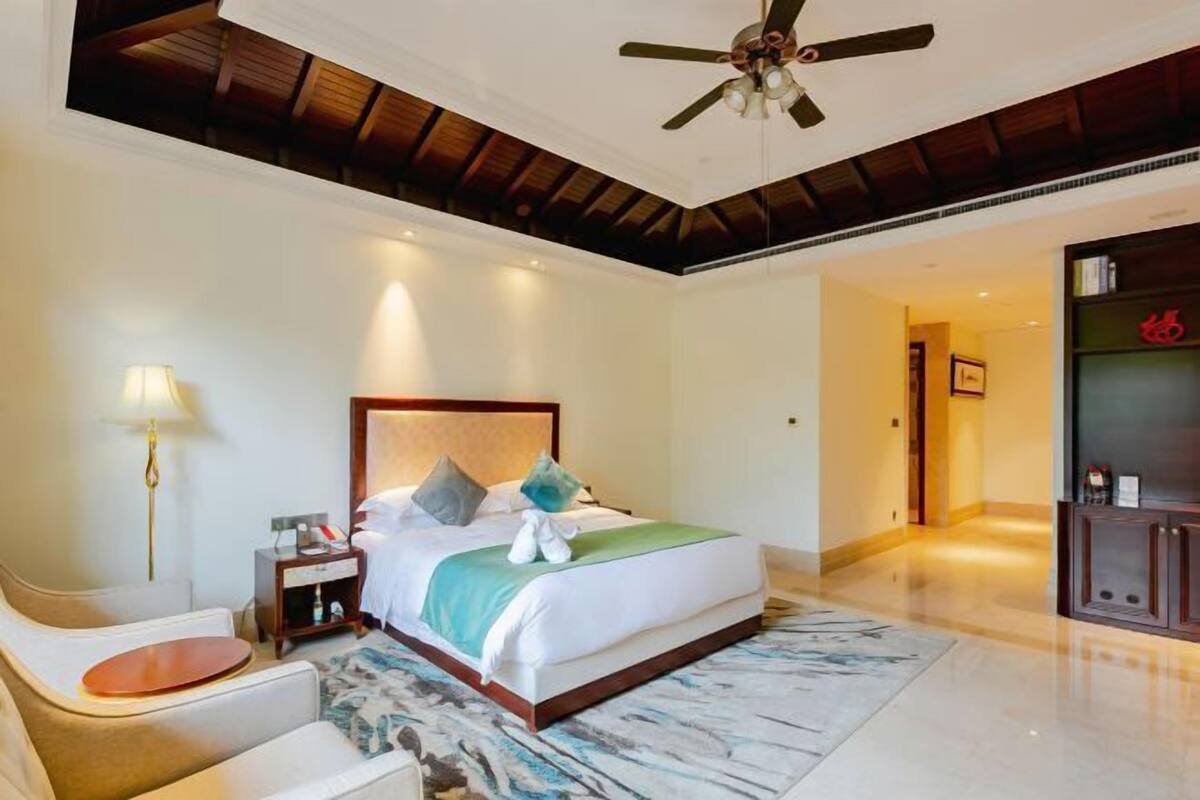 A hotel room with a large bed, a ceiling fan, a nightstand, a lamp, and a view of the hallway.