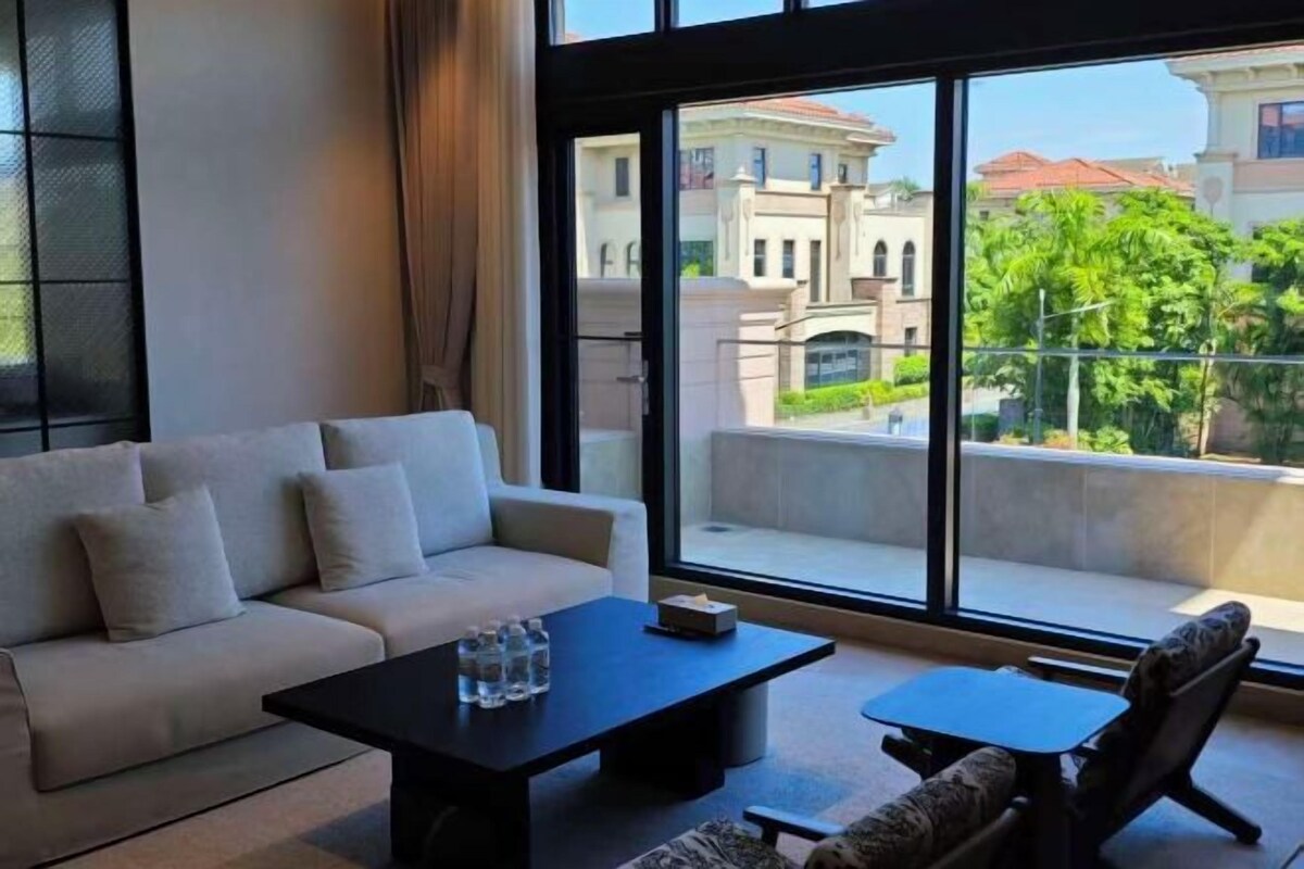 A modern living room with a sofa, coffee table, and armchair, large windows with a view of buildings and trees.
