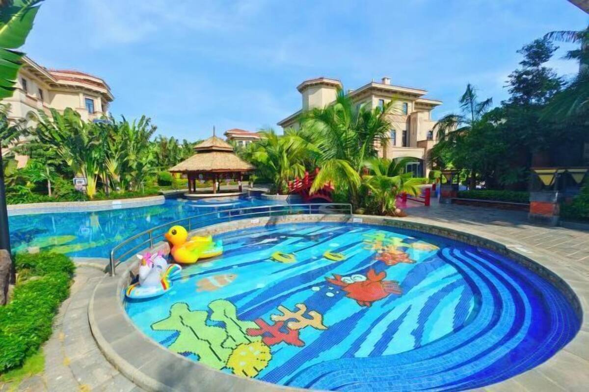 A swimming pool with a colorful mosaic design, surrounded by palm trees and a building with a red roof.