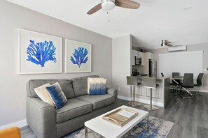 Een moderne woonkamer met een grijze bank, twee blauwe kunstwerken, een witte salontafel, en een eethoek met een bar en stoelen.