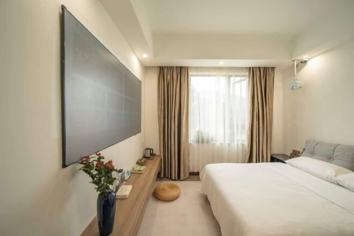 A modern hotel room with a large bed, a flat-screen TV mounted on the wall, a wooden side table with a vase of flowers, and a window with sheer curtains.