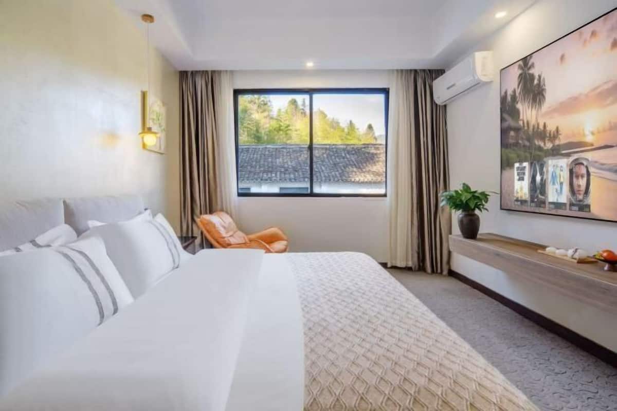 A hotel room with a large bed, a chair, a window with curtains, a plant, and a wall-mounted TV.