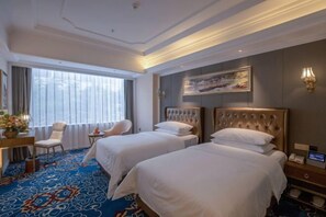 A hotel room with two beds, a desk with a chair, a large window with curtains, and a painting on the wall.