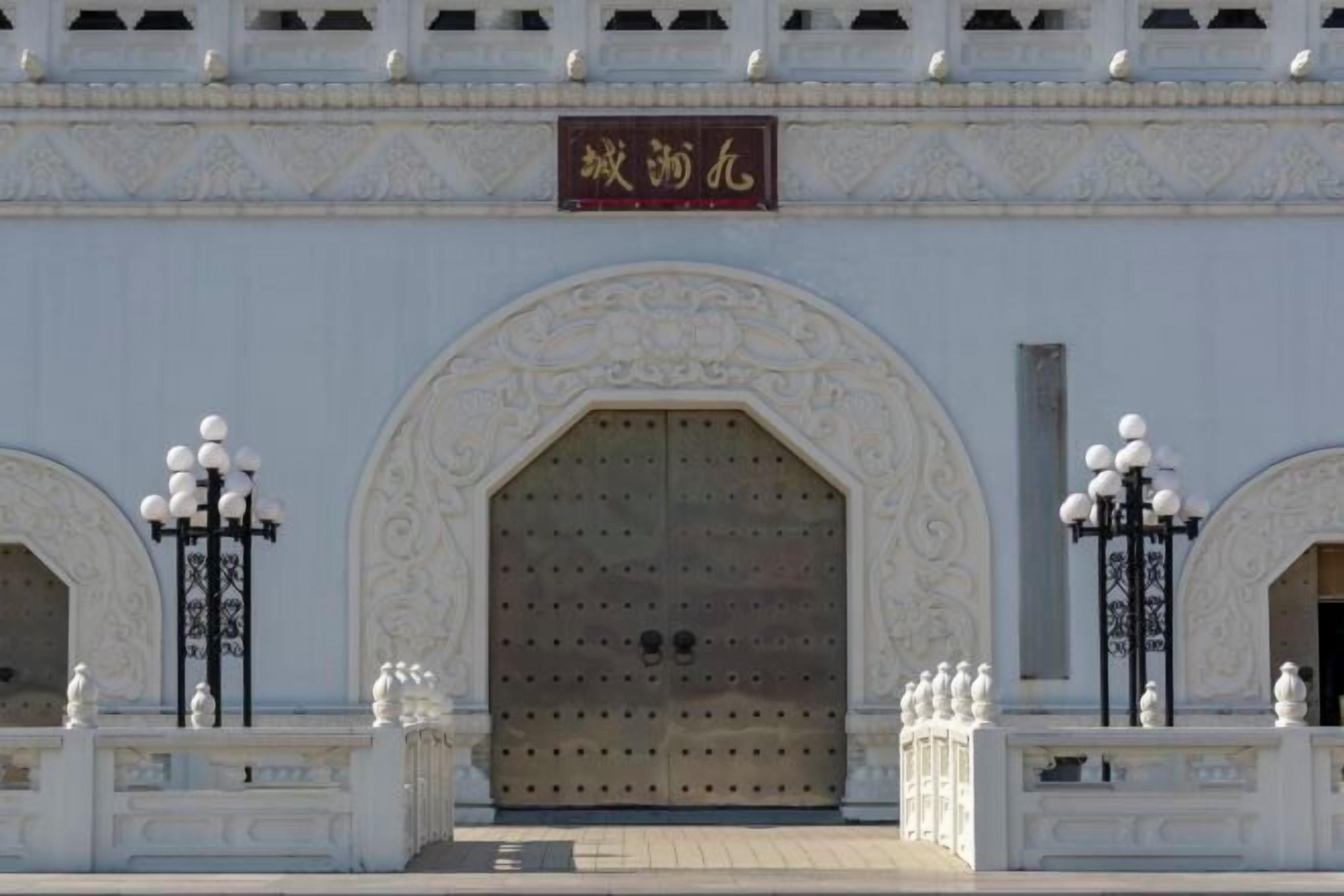 A large, ornate double door with intricate carvings, flanked by two lamp posts with spherical lights, and a sign above the door.
