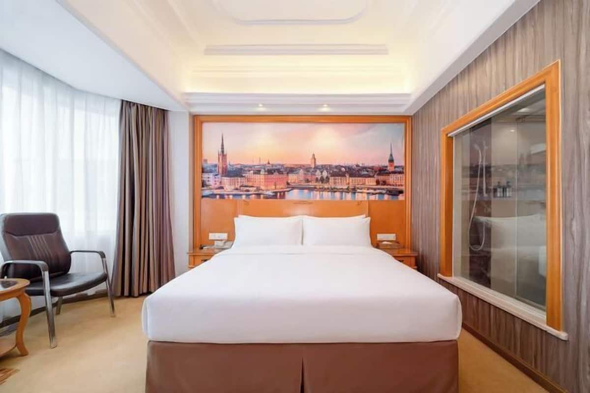 A hotel room with a large bed, a cityscape painting on the wall, a window with a view, and a chair.