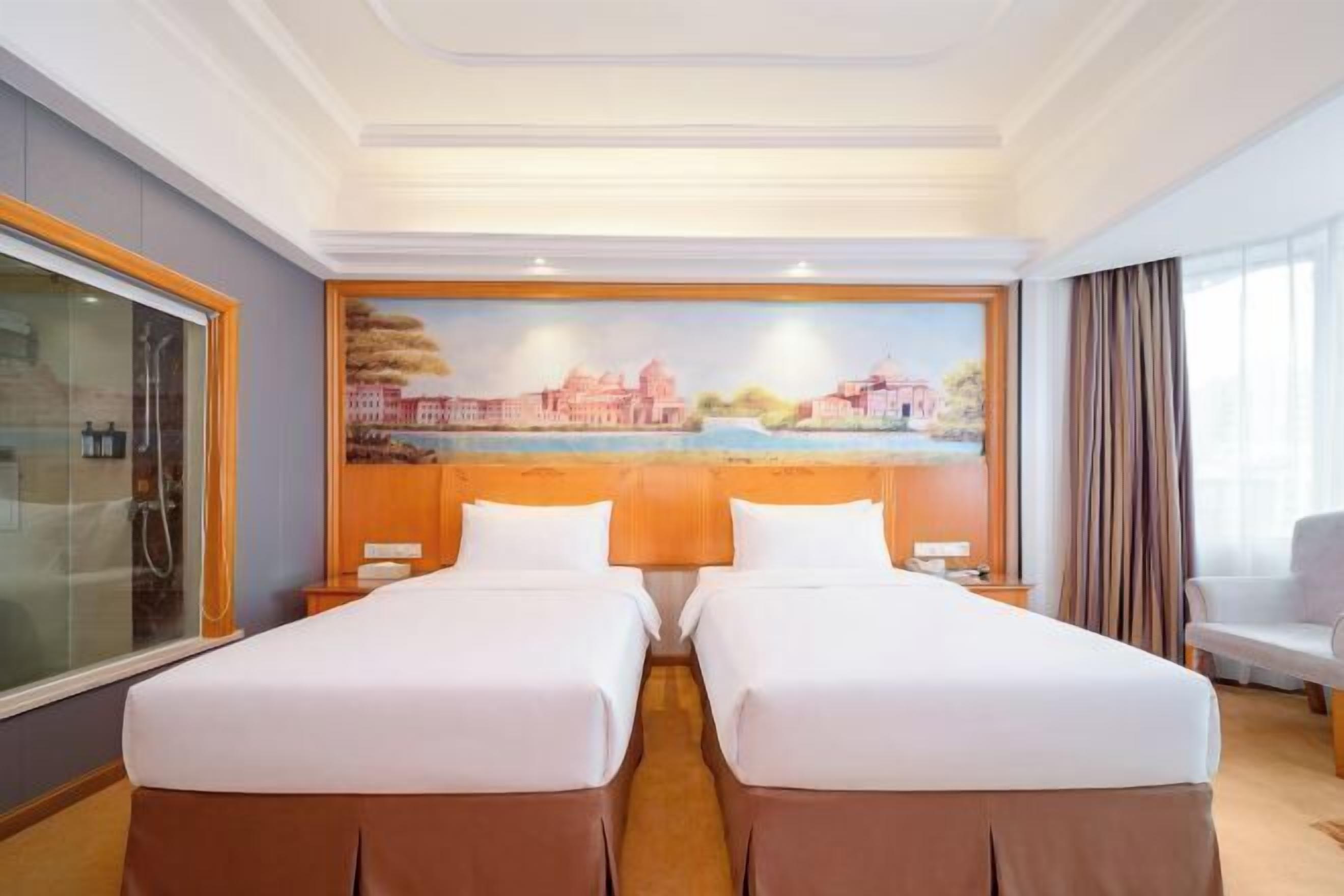 Habitación de hotel con dos camas, un cuadro de un paisaje urbano, una ventana con cortinas y una silla.