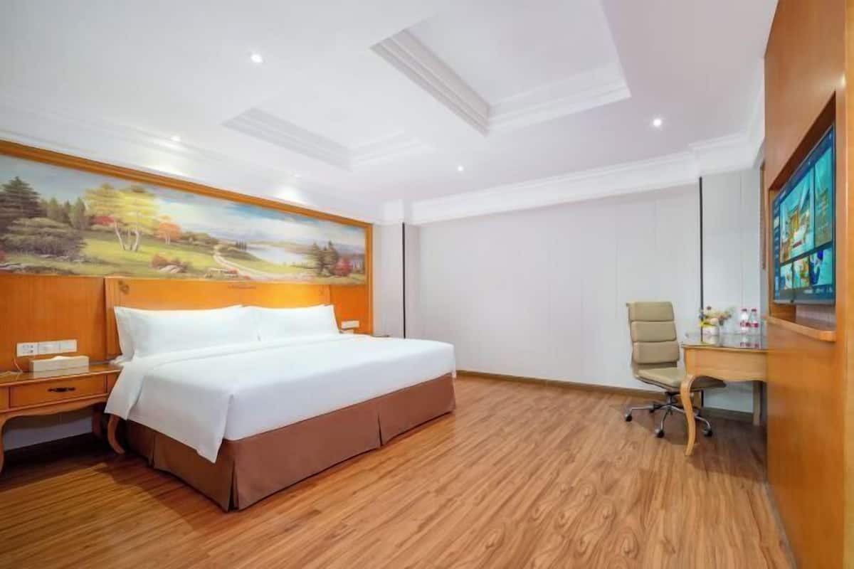 A hotel room with a large bed, a desk, a chair, a painting on the wall, and a TV on the wall.