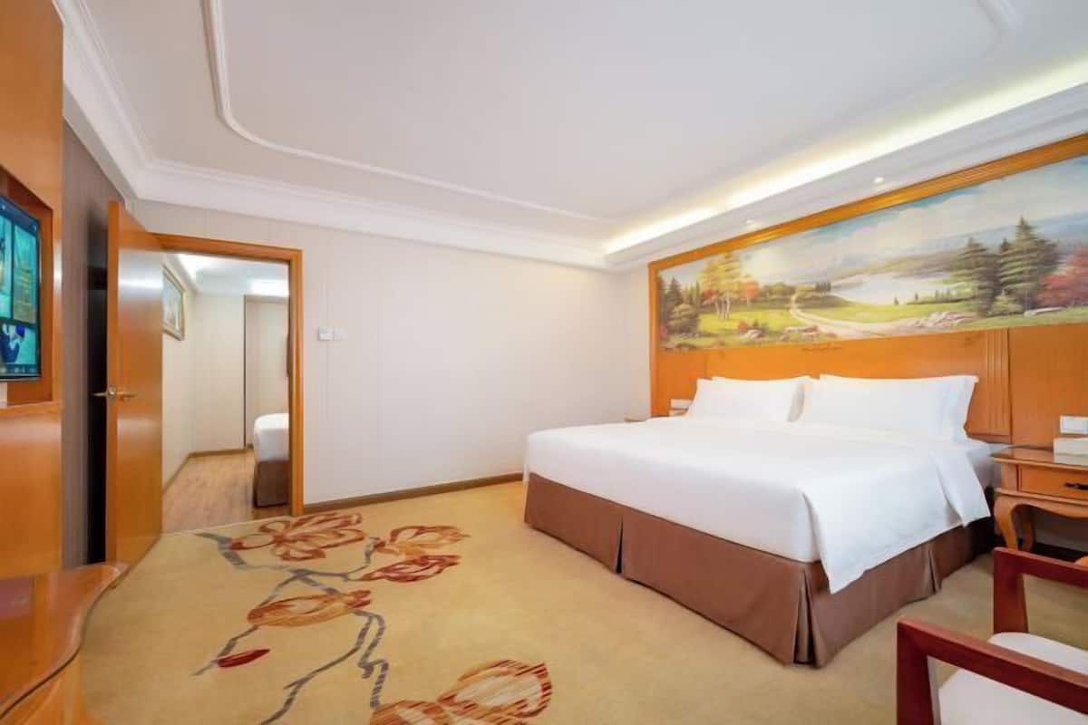 A hotel room with a large bed, a painting on the wall, a wooden desk, and a carpet with a floral pattern.