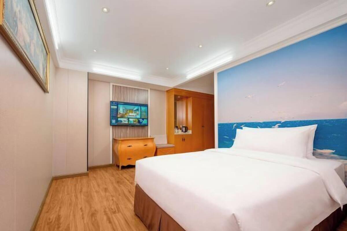 A hotel room with a large bed, a wooden dresser, a television, and a mural of the ocean on the wall.