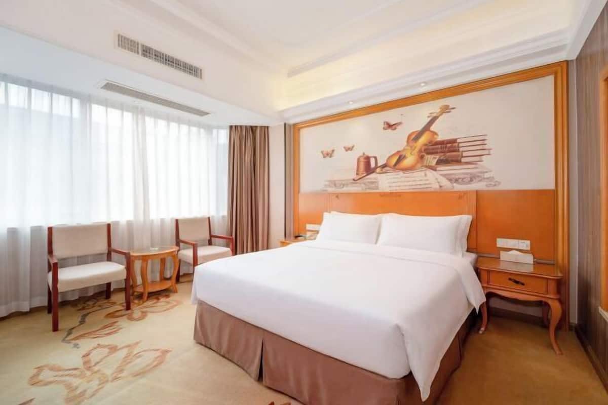 A hotel room with a large bed, a desk, two chairs, a painting on the wall, and a window with curtains.