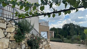 A stone wall with a metal railing, a building with a balcony, and a gravel driveway leading to a forested area. - House with breathtaking views of the Luberon (Caseneuve)