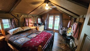 Une chambre confortable située dans les combles, avec un grand lit, un bureau et une chaise. On y trouve une fenêtre avec des rideaux, un ventilateur de plafond et une vue sur l’extérieur.