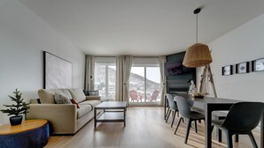 Una sala de estar moderna con sofá, mesa de centro, mesa de comedor y sillas. Hay un ventanal con cortinas, un televisor y una chimenea.