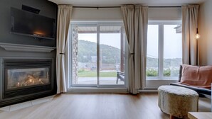A living room with a fireplace, a sofa, a coffee table, and a view of a garden and hills through large windows. - L104-Condo ski in ski out /vélo in vélo out (Bromont)