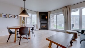 A modern living room with a fireplace, large windows, a dining area, and a sofa. - L104-Condo ski in ski out /vélo in vélo out (Bromont)