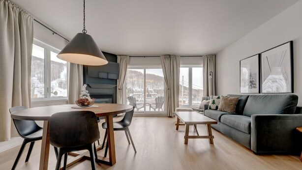 A modern living room with a dining area, large windows, and a view of a snowy landscape. - L104-Condo ski in ski out /vélo in vélo out (Bromont)