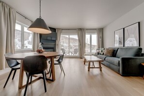 A modern living room with a dining area, large windows, and a view of a snowy landscape. - L104-Condo ski in ski out /vélo in vélo out (Bromont)