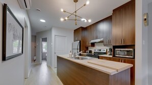 A modern kitchen with wooden cabinets, a central island, stainless steel appliances, and a unique chandelier. - E202-Condo ski in ski out / vélo in vélo out (Bromont)