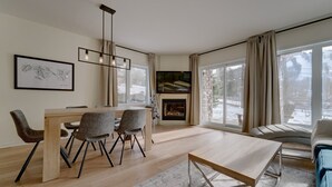 Un salon moderne avec une table à manger en bois, des chaises recouvertes d’un tissu gris, une cheminée et de grandes fenêtres avec des rideaux.