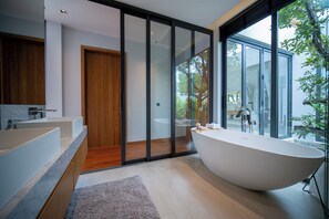A modern bathroom with a freestanding bathtub, a wooden door, and a large window offering a view of the outdoors. - Botanica Foresta 4BR Villa by NLA (Thep Krasattri)
