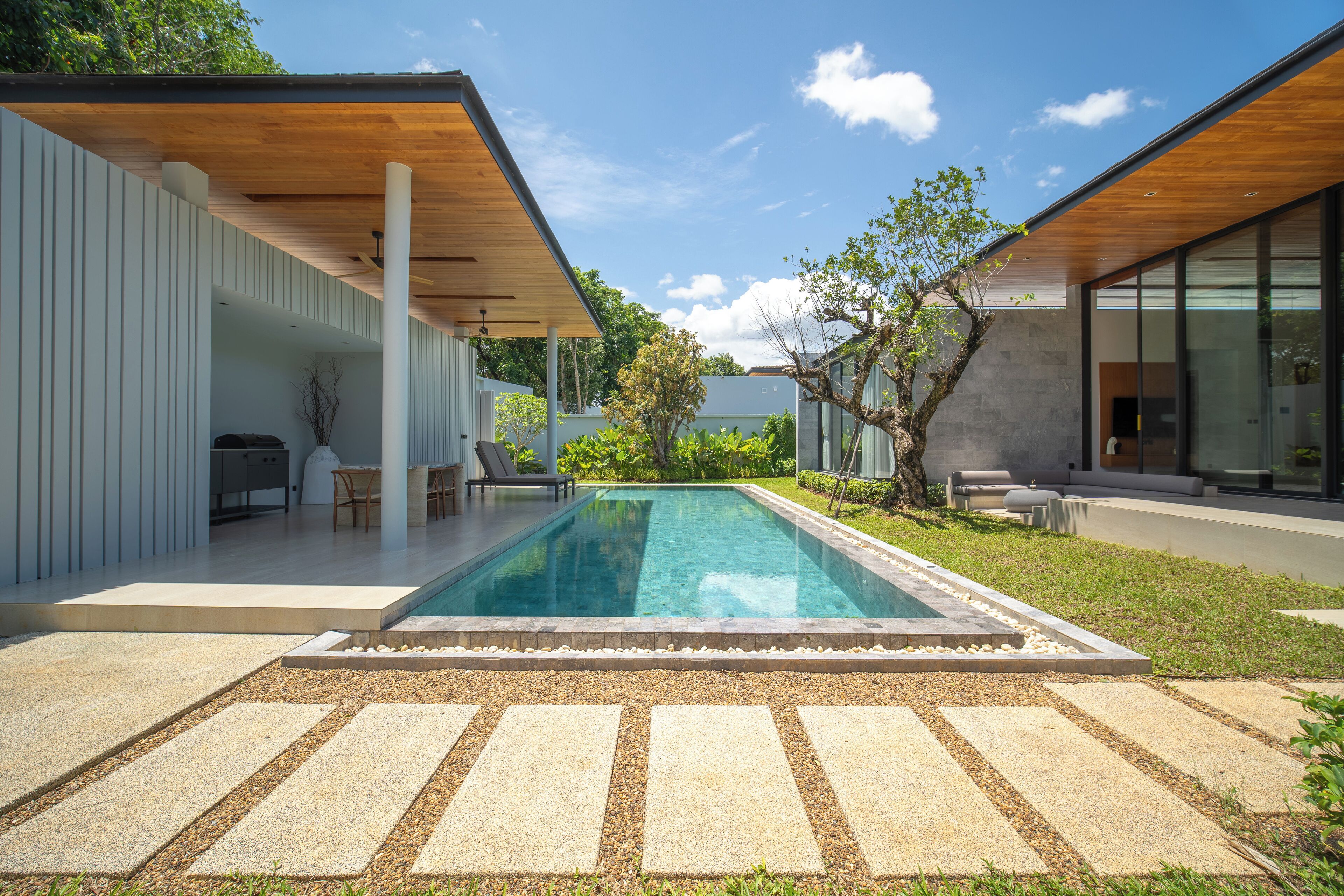 A modern outdoor pool area with a stone deck, a wooden pergola, and a seating area with chairs and a table.