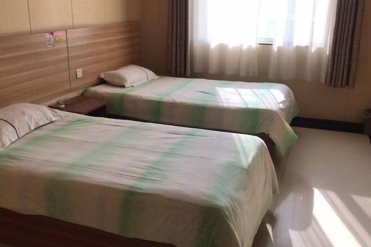 Una habitación con dos camas, cabecero de madera, una ventana con cortinas y una mesita de noche pequeña.