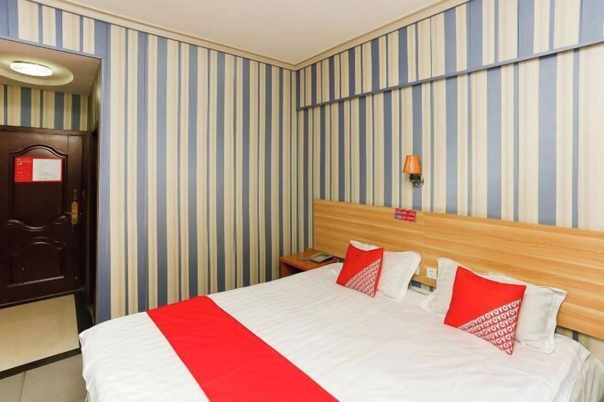 Una habitación de hotel con papel tapiz a rayas, un cabecero de madera, una cama con ropa de cama blanca y roja y una mesita de noche con una lámpara.