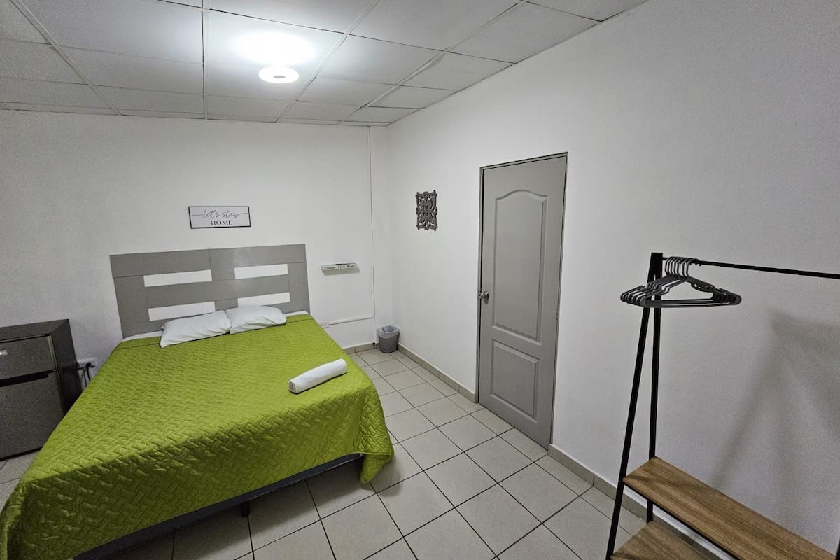 Un dormitorio con cama, un banco, perchero, una puerta y un cubo pequeño.