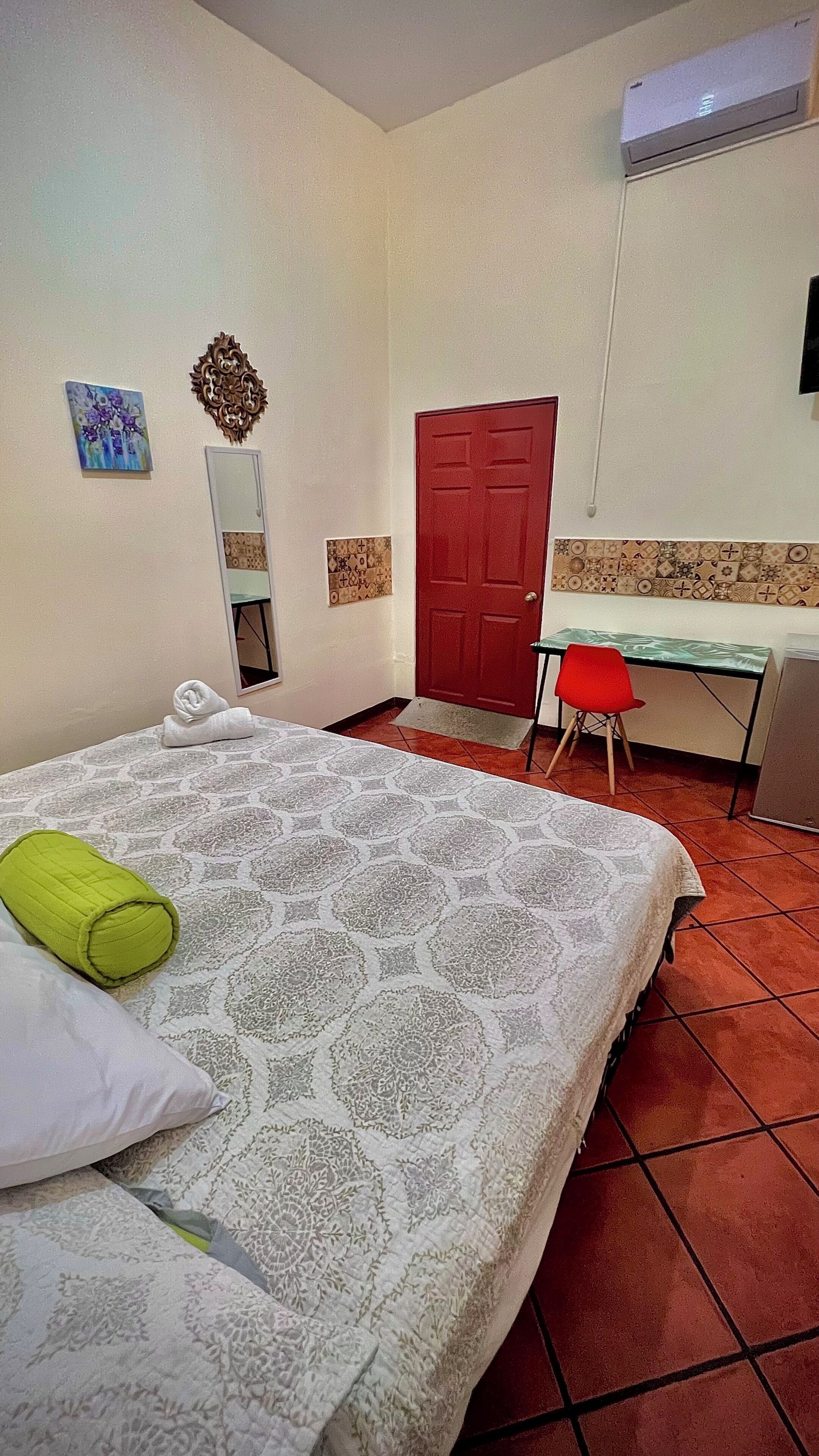 Una camera d'albergo con un letto, una scrivania, una porta rossa e un condizionatore d'aria.