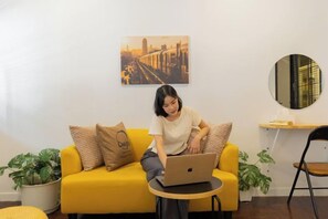 A person sitting on a yellow sofa with a laptop, a round mirror, and a cityscape painting on the wall. - Ombra House No26 (Chiang Mai)